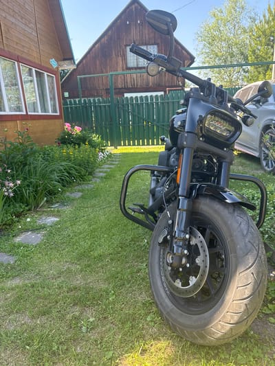 In a residential yard located in the city of Beryozovka, Russia, a black motorcycle is parked on a grassy lawn. The motorcycle, a Harley-Davidson Fat Bob, faces forward, with its front tire prominently displayed. The vehicle features a black frame, handlebars, and a distinctive LED headlight. The background reveals a wooden house with a red trim and white windows, a green wooden fence, and lush greenery, including flowers and trees. A silver car is partially visible behind the motorcycle. The sun casts a warm glow on the scene, suggesting it is daytime.