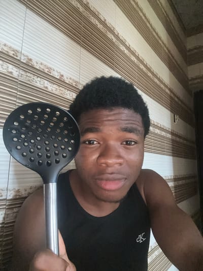 A man is lying on a tiled floor, holding a slotted spoon close to his face. The floor tiles are patterned with alternating stripes of white and brown with a floral design. He is wearing a black t-shirt. The location is Obinze, Nigeria.