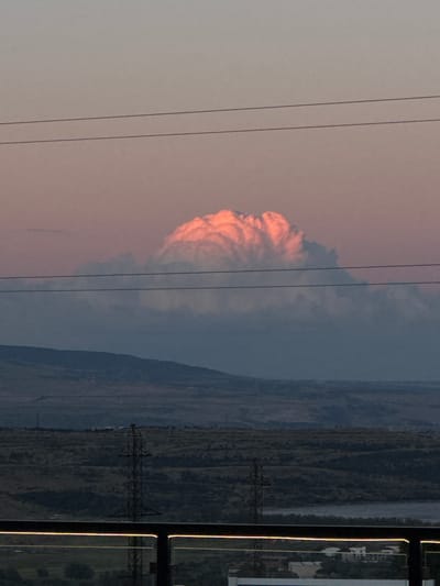 The image depicts an outdoor scene, likely in a rural or semi-rural setting, given the presence of power lines and pylons. The foreground is dominated by three thin, dark power lines running horizontally across the frame, suggesting they are in front of the viewer. To the left, a large, fluffy cloud formation is visible, illuminated by a warm, pinkish-orange light, indicative of either sunrise or sunset. The cloud has a textured, almost sculpted appearance.
The background features a landscape of rolling hills or mountains, covered in dark green vegetation. A body of water, possibly a river or a lake, can be seen in the middle ground, reflecting the muted colors of the sky. Further into the background, a power pylon is visible, a stark industrial element against the natural landscape. On the far right of the image, a building with a white facade and dark window frames is partially visible, suggesting the presence of human habitation or infrastructure. The overall mood is serene and somewhat dramatic due to the striking cloud formation and the natural light. The time of day is either dawn or dusk. No text is visible in the image.