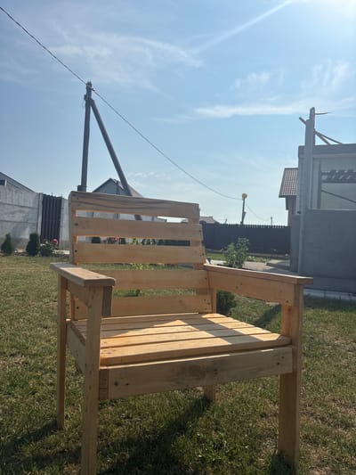 The image shows a light-colored wooden chair placed on a grassy lawn. The chair has a slatted seat and backrest, and armrests. In the background, there is a concrete fence, a dark brown fence, and several houses. A utility pole with wires is visible in the sky. The scene appears to be an outdoor residential area. The location is identified as Skidzieĺ, Belarus.