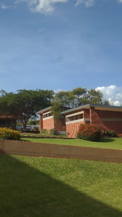 Two brick buildings with white-trimmed windows stand next to a grassy lawn under a bright blue sky with scattered clouds. Green trees partially obscure the buildings. A concrete pathway separates the buildings from the lawn. The lawn is green and well-maintained. The lighting suggests a sunny day.