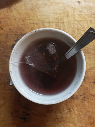 A close-up, overhead shot shows a white ceramic mug filled with dark red tea. A tea bag, made of a mesh material, is submerged in the tea, with its string extending out of the mug and attached to a small white tag. A silver-colored spoon rests inside the mug, partially submerged in the tea and angled towards the right side of the frame. The mug sits on a wooden surface with a visible grain pattern, suggesting it could be a table or countertop. The lighting in the scene is soft and diffused, casting subtle shadows and highlighting the sheen of the tea and spoon. The background is out of focus, emphasizing the mug and its contents. The location is specified as Шарпілаўскі сельскі Савет, Belarus.