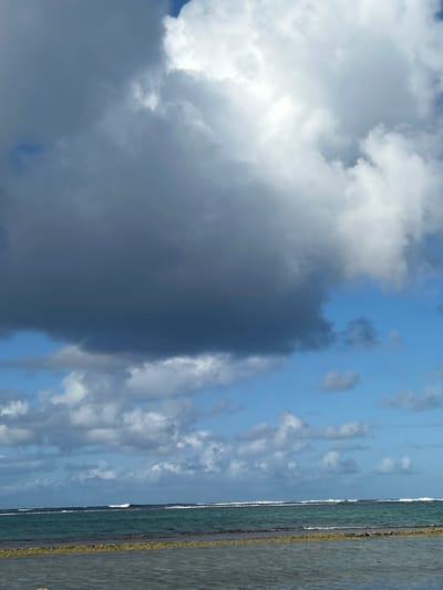 The image displays a coastal landscape in Le Moule, France. The sky occupies the upper two-thirds of the frame, dominated by a large, dark grey cloud mass covering the upper left quadrant. This transitions to brighter white, illuminated clouds in the upper right, where sunlight is evident. Patches of clear blue sky are visible, along with scattered white cumulus clouds, particularly above the horizon line. The lower third of the image features the ocean. The mid-ground water exhibits a deep teal to blue-green color, with several distinct, parallel lines of white-capped waves breaking in the distance. The immediate foreground consists of a shallow, calmer body of water with a greenish-grey hue, separated from the deeper ocean by a band of darker, irregular material, consistent with submerged rock or algae. No human figures, man-made structures, or discernible activity are present.