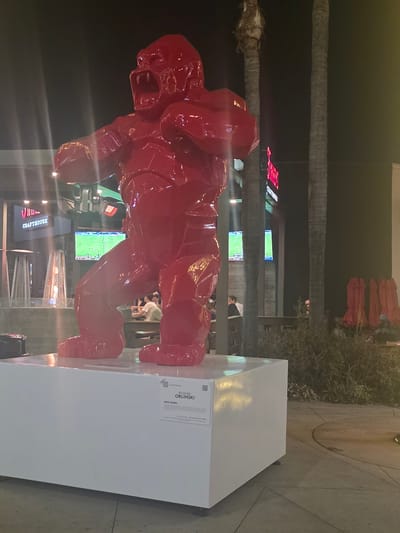 This is a nighttime photograph taken outdoors, likely in the city of Burbank, United States of America, given the visible sign. The primary subject is a large, bright red, stylized sculpture of a gorilla, positioned on a white rectangular pedestal. The sculpture is abstract and angular, with its mouth open in a roaring expression.

In the background, there are palm trees. To the left of the sculpture, a building facade is visible with signage that reads "J.R.'S CRATHOUSE" and "BURBANK." There are also outdoor heaters and what appear to be screens displaying a sports game. A few individuals are seated at tables in the background, partially obscured by the sculpture and foliage.

To the right of the sculpture, more palm trees are present, along with red outdoor umbrellas folded and leaning against a railing. The ground is paved with concrete. A plaque is attached to the white pedestal of the sculpture, bearing text that includes "RICHARD ORLINSKI" and "WILD KONG." The overall scene appears to be an outdoor dining or entertainment area at night.