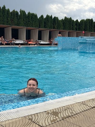 A woman is swimming in a pool, smiling at the camera. She has red hair and is wearing a black swimsuit. The pool is filled with blue water and is surrounded by a tiled border. In the background, there are cabanas and trees. The sky is cloudy. The image was taken in the city of Stara Pazova Municipality, Serbia.