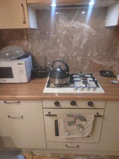 The image shows a kitchen counter with a microwave, a kettle on a gas stove, and a frying pan. The kitchen has tiled walls and wooden cabinets. There are also drawers and an oven visible. The scene appears to be in a residential kitchen. The image does not provide any information about the city of Nalchik, Russia or any specific location within it.