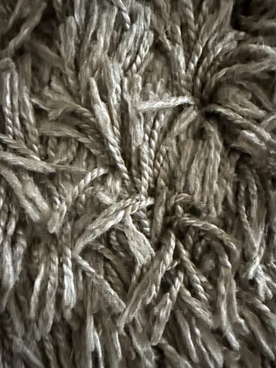 This is a close-up, overhead shot of a shaggy, textured rug. The rug is a neutral grey color and appears to be made of thick, intertwined yarn. The fibers are long and somewhat disheveled, creating a plush and inviting surface. The texture is complex, with individual strands of yarn twisted together to form thicker cords, and these cords are then woven into the overall fabric of the rug. Some areas are denser and more tightly packed, while others are looser, with individual strands fanning out. Light and shadow play across the surface, highlighting the depth and dimension of the rug's pile. There is no text visible in the image. The subject is solely the rug itself, with no people, specific objects, or environmental cues to indicate a particular scene or activity. The close-up perspective and lack of context make it impossible to determine the time of day, weather, or location beyond the general setting of a rug.
