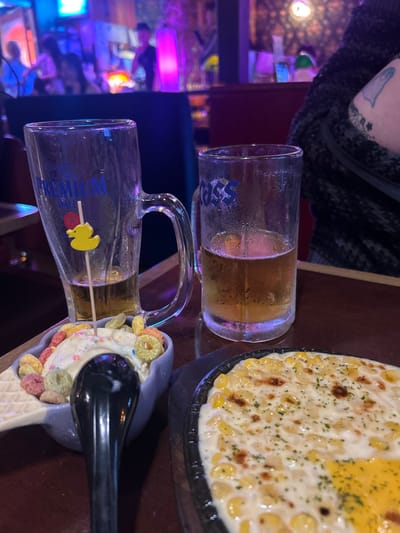 This image captures a dimly lit indoor scene within a bar or pub in 마포구, South Korea. The primary focus is a dark wooden table displaying partially consumed drinks and food. On the left, a large clear glass "Terra Premium" beer mug, nearly empty with a small amount of light-brown liquid and a yellow rubber duck skewer, stands next to a "Cass" branded clear glass beer mug, approximately one-third full of light-brown beer. Both mugs show condensation. To the left of the Cass mug, a light blue ceramic bowl shaped like a waffle cone holds vanilla ice cream topped with colorful ring-shaped cereal and sprinkles, with a black spoon resting in it. Adjacent to this, on a black sizzling plate, is a dish of baked corn kernels in a creamy white sauce, garnished with green herbs, and featuring a section of melted yellow cheese or sauce on the lower right. On the far right, the upper arm and shoulder of an individual wearing a dark, textured garment are visible, marked by a light blue ghost-shaped tattoo. In the blurred background, several figures, likely patrons, are present amidst ambient blue and purple lighting, with one figure near a prominent pink glowing pillar. The scene depicts a social gathering for drinking and dining.