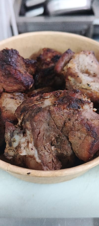In Ploske, Ukraine, a close-up, eye-level shot captures a bowl filled with cooked meat. The meat appears to be pork, with visible layers of fat and muscle tissue, and a dark, seared exterior indicative of grilling or roasting. The bowl itself is a light brown, disposable container, likely made of paper or cardboard, with a slightly worn edge. The background is blurred, showing a metallic surface and an out-of-focus kitchen environment. The lighting is natural and comes from above, highlighting the texture of the meat.