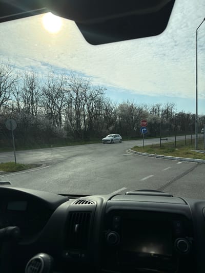 The image depicts a daytime scene from the interior of a vehicle, looking forward through the windshield at an asphalt road intersection in Zagore, Bulgaria. The sun, high in the sky and partially obscured by the top edge of the windshield, causes a bright glare. The sky is largely covered by thin, textured clouds, with some patches of blue.
In the foreground, the dashboard of the viewer's vehicle is visible, showing a dark gray instrument cluster, air vents, and a manual gear shift knob. The vehicle is positioned at an intersection with white dashed road markings on the asphalt.
To the left, a gray circular sign (backside visible) stands near the roadside. Beyond the road, a dense line of deciduous trees, many with white blossoms, indicates late winter or early spring. Green grass grows along the road edges and in a small, curved median at the intersection.
To the right, a prominent red octagonal "STOP" sign is mounted on a pole, with a blue circular sign below it displaying a white curved arrow pointing right, indicating a mandatory right turn. Further right, a tall, slender streetlight pole extends vertically, and a dark wire fence separates the road from an adjacent grassy area. A second vehicle, a light gray two-door hatchback, is visible approaching or waiting at the intersection from the left. A faint outline of another vehicle is barely discernible behind the fence on the far right.