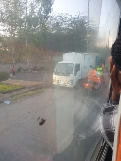 A white box truck parked on the side of a road next to trees. A person in an orange vest stands near the front of the truck, and another person in a yellow vest is nearby. The image is taken through a dirty or foggy window. Part of a person wearing a gray and white plaid shirt is visible in the upper right corner, and a black knit hat is in the upper left corner.