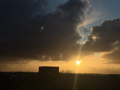The image depicts an outdoor panoramic view of Naaldwijk, Netherlands, at either sunrise or sunset. The scene is dominated by a dramatic sky with heavy, dark clouds in the upper left and center. A bright, low sun is visible through a break in the clouds on the right side, emitting golden light and prominent vertical lens flares. Patches of blue sky are visible above and around the sun.
In the midground, a large, dark, rectangular building, likely commercial or industrial, forms a prominent silhouette, slightly to the left of the image center. A faint rectangular lighter area, possibly a window, is discernible on its front face. To the right of this building, a cluster of bare tree branches is silhouetted against the bright horizon. Below the main building and extending across the bottom of the frame is a darker, expansive, flat structure or ground area.
The distant horizon features a silhouetted cityscape or town outline, including a distinct church spire towards the right of the distant skyline and several communication towers on the far left. A row of vehicles and several streetlights are discernible as silhouettes in the midground, below the distant horizon. A circular green lens flare is present in the upper left quadrant of the image, within the dark cloud formation. No individuals or explicit actions are depicted.