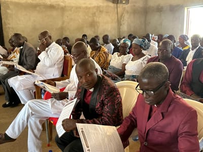 This image depicts a gathering of people, likely attending a religious service, inside a building with concrete walls and a pale ceiling. The attendees are seated in chairs, many of whom are holding and reading documents, suggesting they are following a program or sermon.

In the foreground, two men are prominently featured. The man on the left wears a black suit with a white clerical collar and a patterned dark red and black jacket. He is looking towards the camera with a serious expression and holding a document. To his right, a man with graying hair wears a burgundy blazer over a white clerical collar and is looking down at a document.

Behind them, other individuals are seated. Many are wearing traditional African attire, including brightly colored patterns, as well as Western-style clothing. Several women are wearing headscarves. The overall atmosphere appears somber and focused. The presence of a speaker or amplifier mounted on the wall suggests a formal event.

The image description indicates this scene is located in the city of Kufana, Nigeria. The environment suggests an indoor gathering space within a community, possibly a church or community hall. The lighting appears to be a mix of natural light from an open doorway or window and potentially artificial lighting.