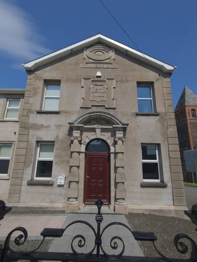 The image shows the front facade of the Masonic Hall in Magherafelt, United Kingdom, on a clear, sunny day. The building, constructed of light-colored stone, features a prominent arched doorway with a dark wooden door and a small white mailbox attached to the wall to the left of the entrance. Above the door, a decorative crest is visible with the inscription "MASONIC HALL" and an eye symbol. To the right of the main building, a section of a red brick tower with arched windows can be seen, characteristic of the nearby tower. A black ornate metal fence is in the foreground, partially obscuring the paved walkway leading to the building. The sky above is a vivid blue with a few wispy white clouds.