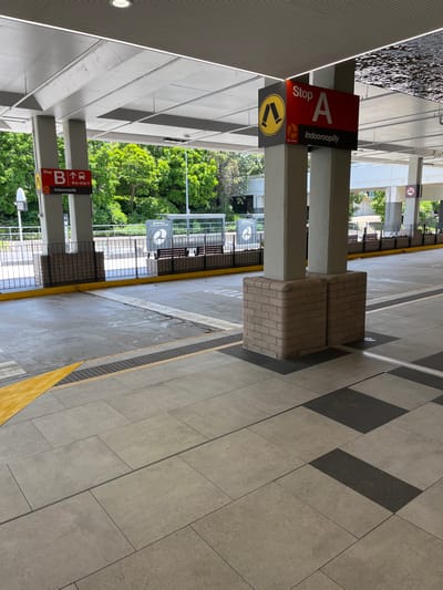 A bright, sunlit outdoor public transport platform, likely a bus stop, is depicted, rotated 90 degrees counter-clockwise. The platform is paved with light grey tiles, some featuring a contrasting dark grey rectangular pattern. A yellow tactile warning strip runs along the edge of the platform.

Two substantial concrete pillars stand on the platform. The left pillar (when viewing the rotated image) features a red and black sign marked "Stop A Indooroopilly", with a yellow circular logo containing a stylized arrow or 'v' symbol below. The right pillar also has a red and black sign, partially visible, which reads "Stop B" and "bus stop C", also followed by "Indooroopilly" and a similar yellow circular logo. Two block-like seats made of light-colored bricks are positioned next to one of the pillars.

In the background, lush green trees and foliage are visible beyond a metal fence, suggesting an open, possibly suburban setting. Further back, another circular sign with a 'C' and a logo can be seen. A "No Smoking" symbol is also visible on a distant pillar. The area appears clean and well-maintained, with no people present, indicating a quiet moment at the Indooroopilly, Australia location. The bright lighting suggests it is daytime, likely a clear and sunny day.