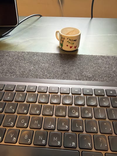 An overhead view captures a workspace, primarily featuring a laptop keyboard and a ceramic mug, resting on a surface partly covered by a grey textured mat.

The main subjects are a dark grey laptop keyboard from the brand "Logi" (Logitech) and an off-white cylindrical ceramic mug with a handle. The keyboard features a bilingual layout with both Latin and Hebrew characters, indicating its use in a region like Kafr Yasif, Israel. Several function keys (F1-F12) are visible, along with alphanumeric keys, punctuation, and symbols. The mug has brown residue inside, suggesting it recently held a beverage like coffee or tea.

On the side of the mug, there is a cartoon drawing of a reclining figure and the Italian text "L'ULTIMO PASSO" (meaning "the last step" or "the final stride"), accompanied by three red heart symbols.

The objects are placed on a dual-textured surface; the laptop sits on the grey textured mat, while the mug is on an adjacent lighter, smoother surface, possibly a desk. Part of a black electronic device (likely a smartphone or another portable device) and a black cable are visible in the bottom left corner, indicating a busy work setup. There are no people visible. The lighting appears to be ambient indoor light.