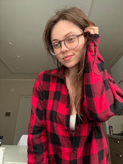 A young woman with brown hair and round glasses poses while wearing a red and black flannel shirt. She has a slight smile and is holding her hand near her head. A hint of a white top can be seen underneath the open flannel. The background consists of a plain light gray wall with some visible fixtures. The image is taken at an angle.
