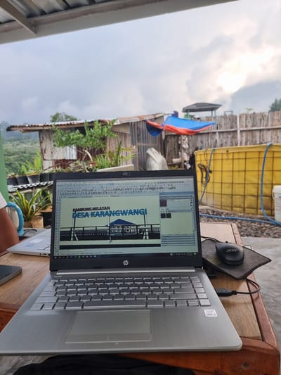 The image shows a laptop displaying a SketchUp Pro 2021 file titled "Kampung Nelayan Desa Karangwangi," suggesting a design or planning document for a fishing village in Karangwangi. The laptop is on a wooden table outdoors, with an Acer mouse nearby. The background reveals a rural scene with buildings, vegetation, and a view of mountains, indicating the location is likely in or near Sindangjaya, Indonesia, based on the file name context. There are also a yellow container and other objects in the background.