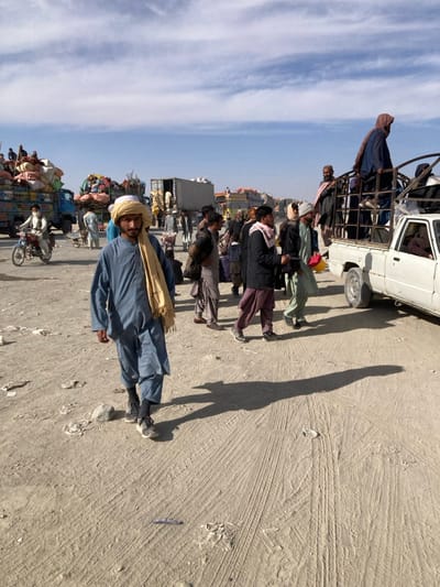 The image depicts a busy, dusty, unpaved area in Chaman City Tehsil, Pakistan, under a bright sky with scattered clouds. Numerous individuals, predominantly adult males in traditional attire (shalwar kameez, turbans, shawls), are present.
In the foreground, a man wearing a blue shalwar kameez and a light-colored turban and shawl walks forward, casting a long shadow to his right. The ground exhibits tire tracks and scattered debris.
In the midground and background, multiple vehicles are visible. To the right, a white pickup truck with an open metal cage in its bed is occupied by several passengers, some standing. To the left and in the background, a line of large, heavily overloaded trucks, stacked high with various goods such as sacks and bales, extends into the distance. A man on a red motorcycle passes by these trucks on the left. People are seen walking and congregating around the vehicles throughout the scene. The lighting indicates strong daylight, with long shadows suggesting morning or late afternoon.