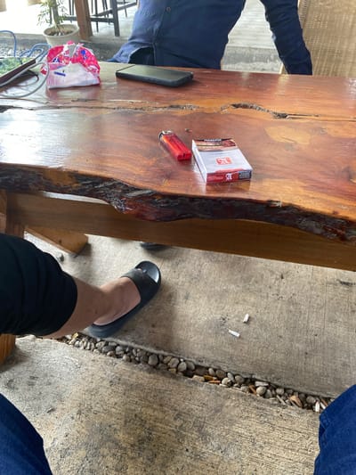 The image shows a close-up of a rustic wooden table with several items on its surface. A red cigarette lighter and a pack of Sampoerna cigarettes are prominently displayed. To the left, a smartphone and a crumpled pink and white package are also visible. In the foreground, a person's legs are visible, wearing black shorts and black flip-flops. They are seated at the table, with one foot resting on the concrete ground. The environment appears to be an outdoor seating area, possibly a cafe or restaurant, with a concrete floor and a row of small stones separating the seating area from a pathway. Several cigarette butts are scattered on the ground. The background shows a blurry view of chairs and possibly other tables, suggesting a social setting. The location is identified as Aluepiet, Indonesia.