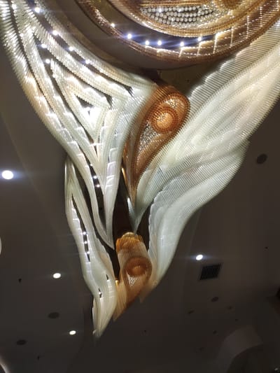 The image shows a large, ornate chandelier hanging from a ceiling. The chandelier is made of numerous transparent, elongated elements arranged in a cascading, wave-like pattern. Some sections of the chandelier are illuminated by embedded lights, creating a bright, shimmering effect. There are also larger, spherical lights on the ceiling. The ceiling itself is a dark, smooth surface with several recessed lights and ventilation grates visible. The chandelier has a distinctive, flowing design with metallic accents in gold and silver tones. The overall impression is one of grandeur and elaborate decoration. No people are visible in the image. The location is identified as Ürümqi, China.