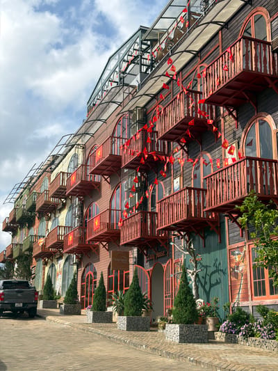 The image shows the exterior of a building in Măng Đen, Vietnam. The building has multiple levels with a distinctive architectural style, featuring red balconies decorated with Vietnamese flags. The facade incorporates wood and other materials, creating a unique aesthetic.

A sidewalk is visible in the foreground, along with parked cars. The sky above is partly cloudy. Lush greenery surrounds the building.