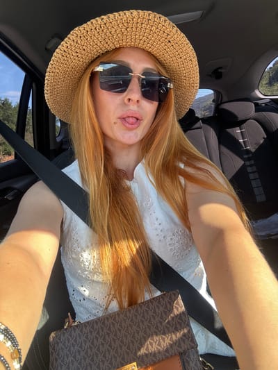 A Caucasian female with long, auburn hair is captured in a selfie taken from inside a vehicle, likely in the city of Prijeradi, Montenegro. She is wearing a straw hat and large, dark-lensed sunglasses, and has her tongue sticking out. Her attire includes a white, eyelet-detailed top, and a brown and tan Michael Kors monogrammed purse is visible. A black seatbelt is fastened across her chest. The background shows the interior of the car and a glimpse of a mountainous, wooded landscape through the window, suggesting travel. The lighting indicates it is daytime, with sunlight casting shadows.