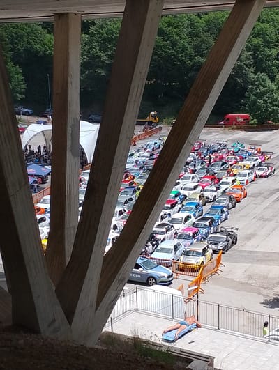 The image depicts an outdoor event, possibly a race or car show, set in a large paved area. In the foreground, large concrete pillars from a bridge or overpass dominate the view, casting shadows and framing the scene. Beyond the pillars, a multitude of colorful cars, many of them appear to be modified or racing vehicles with spoilers and racing decals, are parked in neat rows. Some of the cars are white, red, blue, yellow, and black, with various sponsor logos and designs.
There are also several people visible. A person is lying on their back on a blue mat on a tiled area, with a black mobile phone or remote beside them. Further away, near the cars, several individuals are standing and observing the vehicles. There's also a police car with "POLIZIA" written on it. Tents or canopies are set up in the background, suggesting a service or spectator area. The environment is characterized by a bright, sunny day, with lush green trees forming a backdrop behind the event area.
The caption specifies the location as the city of Rieti, Italy. Given the landscape and the presence of what appears to be a racing event or car gathering, this scene could be taking place at a venue like the Autodromo di Rieti or a similar open area within or on the outskirts of the city. The overpass structure in the foreground is a distinctive architectural element.