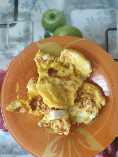 The image shows a plate of scrambled eggs. The plate is orange and has a floral pattern. The background is a countertop with a floral and apple design. The image was taken in Askiz, Russia.