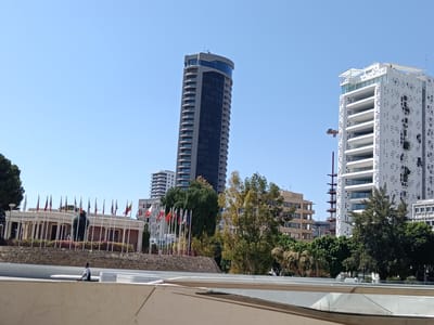 This is a daytime, outdoor photograph taken on a clear, sunny day in Nicosia, Cyprus. The image captures a city skyline with several modern buildings and a row of international flags.

In the foreground, a light-colored, curved structure dominates the bottom of the frame, suggesting an elevated walkway or viewing area. A single person, a man wearing a white shirt and dark shorts, is visible walking along this structure.

The middle ground features a low, classical-style building with columns, fronted by a line of approximately twenty flags representing various nations. Behind this building, a tall, dark, cylindrical skyscraper with many balconies rises against the bright blue sky. To the right of this skyscraper, a striking white, geometric modern building with a distinctive pattern of square openings is visible. A construction crane, colored orange and red, is also present, positioned near the white building. Further in the background, other buildings of varying heights and architectural styles are present, contributing to the urban landscape. Lush green trees are scattered throughout the scene, softening the architectural elements.

The overall impression is one of a modern, international city on a pleasant, clear day. The presence of numerous flags suggests a location of diplomatic or international significance. The time of day is clearly indicated by the bright sunlight and the clear, cloudless sky.
