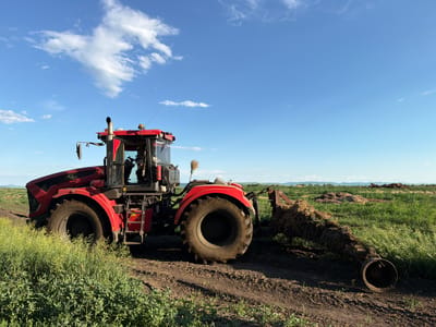 A red tractor is parked in a field under a bright blue sky with scattered white clouds. The tractor is equipped with large tires and appears to be engaged in agricultural work, as indicated by the tilling attachment loaded with organic matter at the rear. The field is covered with green vegetation, and in the distance, a low mountain range is visible. The scene suggests a rural setting, likely in or near Zelenyi Dol, Russia.