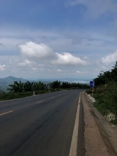 The image captures a scenic view of a curved asphalt road winding through a lush, mountainous landscape. The road, marked with yellow and white lines, is flanked by a grassy shoulder and dense vegetation, including banana trees and tall grasses. In the distance, rolling hills and a body of water are visible under a partly cloudy sky. A blue traffic sign with white symbols, indicating a lane split and a recommended route, stands on the right side of the road. The overall scene suggests a quiet, possibly remote, location, consistent with the mountainous terrain found around Phan Son, Vietnam. There are no people or animals present in the image.