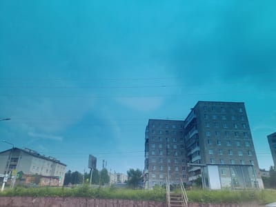 The image depicts a residential area in Chusovoy, Russia, characterized by Soviet-era apartment buildings. In the foreground, a stone retaining wall runs horizontally, topped with green grass and vegetation. A rusted metal staircase is visible on the right. Behind the wall, several apartment buildings are present. The most prominent building on the right is a tall, multi-story concrete structure with numerous windows. To its left, a smaller, lighter-colored apartment building with a more angled facade is visible, featuring several balconies and a smaller building in front of it with green and brown brickwork. In the background, more apartment buildings and trees can be seen, along with utility poles and wires crisscrossing the sky. The sky is a vibrant, uniform turquoise, suggesting either a filtered lens or a highly saturated sky. A billboard or signpost is also present on the left side of the frame. The scene appears to be an outdoor, daytime view of a typical urban landscape in Chusovoy.