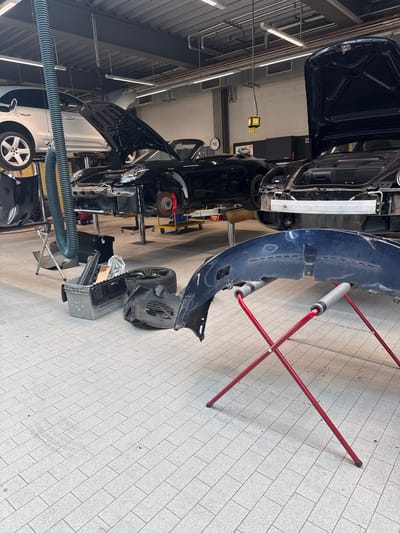 The image depicts a repair shop interior in Hofheim am Taunus, Germany. Several cars are undergoing maintenance, with parts removed and placed on the floor. A blue car bumper rests on a red support stand. A black car with its hood open is elevated on a lift. A silver car is visible in the background. Various tools and equipment are scattered throughout the workshop. The floor is tiled.