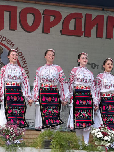 Four young women in traditional Bulgarian folk costumes are standing on a stage, holding hands. Their outfits consist of white embroidered blouses with red trim, black aprons with colorful embroidered patterns, and headbands with white ornamentation. They are smiling and appear to be performing or posing for a photograph. In the background, a large banner with red lettering is visible, partially obscured by the women. The stage is decorated with floral arrangements, including a bouquet of purple flowers on the left and white and red flowers on the right. Green foliage is also present in the foreground. The overall scene suggests a cultural event or festival taking place in Pordim, Bulgaria.
