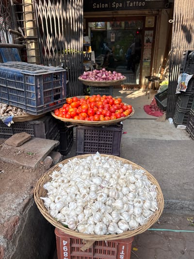 The image shows a street-side vegetable stall in Ambernath, India. Three wicker baskets are arranged on top of stacked plastic crates. The baskets contain, from left to right: red onions, tomatoes, and garlic. The stall is situated in front of a storefront with a partially closed metal shutter. A sign on the storefront reads "Salon Spa Tattoo." The street surface is concrete and asphalt. The environment appears to be an open-air market.
