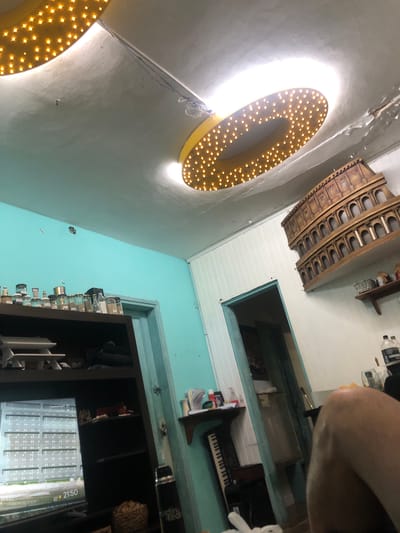 The image shows an interior shot in what appears to be a room in Beccar, Argentina. The walls are painted a light turquoise color with a dark brown bookcase against one. Several glass bottles are mounted along the wall next to the bookcase. A white-painted ceiling features a wooden model of the Colosseum and a rectangular opening to another room. Arched light fixtures with numerous bulbs are mounted on the wall. A small electronic keyboard sits on a shelf in the adjacent room. A portion of a person’s arm is visible in the upper right corner.