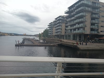 The image shows a view of the Drammenselva river in Drammen, Norway. In the foreground, a white railing is visible. The river occupies the mid-ground, with a wooden dock featuring several moored boats on the left side. In the background, a modern apartment building with glass balconies lines the riverbank. Further in the distance, a bridge spans the river, with buildings and hills visible on the opposite bank. The sky is overcast. Several people are walking on the promenade alongside the building.
