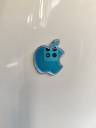 The image displays a close-up of a teal-colored apple logo sticker with a reflective silver outline. Inside the apple shape, there is a graphic representation of a multi-lens camera system, mimicking the design of smartphone cameras. The sticker is placed on a light-colored, possibly laminated or polished surface, which reflects some light, creating soft, elongated highlights. The background is plain and out of focus, suggesting the sticker is the primary subject of the photograph. There is no text visible in the image. The lighting appears to be diffused, potentially from overhead or ambient sources. The setting is not clearly identifiable beyond being an indoor surface.