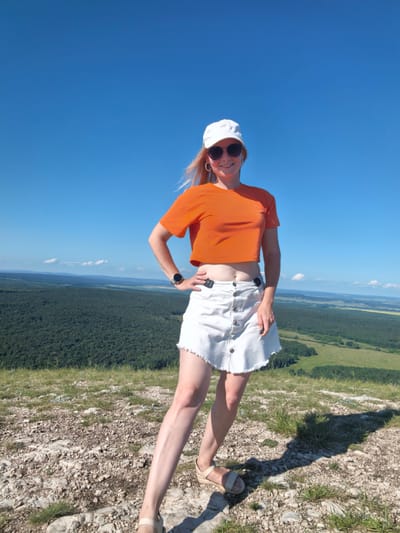 A woman is standing on a rocky, grassy hilltop with a vast green forest and rolling hills stretching out behind her under a clear blue sky. She is wearing a white baseball cap, black sunglasses, and an orange crop top with a white denim skirt that has a frayed hem. Her left hand is on her hip, and her right arm is resting on her skirt. She is wearing beige sandals and a watch on her left wrist. The location is the Shikhan Mountains in Russia, a natural landmark known for its unique geological formations and scenic views.