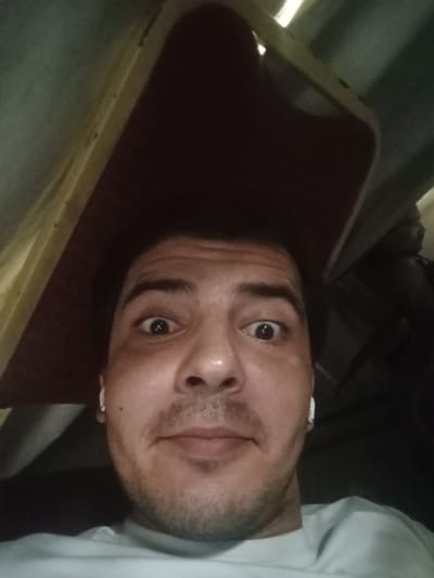 The image is a close-up, low-angle selfie of a man, likely taken indoors. The man has short dark hair, brown eyes, and stubble. He is wearing a white t-shirt and white earbuds. The background is dark and appears to be the interior of a room, with a textured ceiling or overhead structure visible. There are no discernable landmarks or context that would indicate a specific location within Sharjah, United Arab Emirates. The lighting suggests artificial indoor lighting.