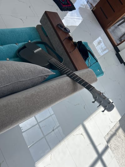 A guitar is placed on a sofa in a home in Kadewatta, Sri Lanka. The guitar is black and gray, with a black body and a gray neck and headstock. The sofa is blue and gray, and it is covered with a patterned rug. The floor is tiled with white and gray tiles, and there are shadows on the floor from the window. The scene is well-lit and the objects are in focus.