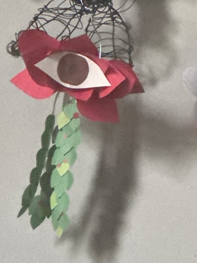 A handmade paper flower, predominantly red with a white and brown center, rests on a gray surface. Attached to the flower is a green paper branch-like structure. Adjacent to the flower is a dark, twisted wire form. A diffuse shadow lies on the gray background. There is no information available in the image to determine if the photo was taken in Winchester, United States of America.