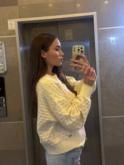 A woman in an elevator holds a phone up to a mirror. She wears a cream-colored, textured sweater, blue jeans, and a light-colored shirt underneath. Her long brown hair falls to her waist. The phone has a decorative case with a black and white design. The elevator has grey walls and a control panel at the bottom of the frame.