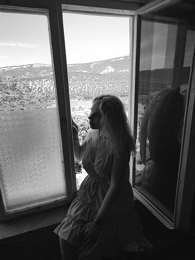 The image depicts a black and white, eye-level indoor scene featuring a woman looking out a window in Терновский округ, Ukraine. A woman with long, light, wavy hair is positioned on the right side of the frame, facing left and looking out a multi-pane window. She is kneeling or sitting on the floor by the window, wearing a light-colored, V-neck, sleeveless dress with ruffled tiers. A necklace with a pendant is visible, and a bracelet adorns her left wrist, with a watch on her right wrist. Her left hand is near the window frame. The primary light source originates from outside the window, creating a backlit effect that casts portions of the woman's right side into shadow.
The window structure consists of multiple panes with weathered light-colored wooden frames. The left window pane's lower section features textured, wavy glass, while its upper section is clear. The right window pane is clear, reflecting a dark, distorted image of the woman's profile and hair. Through the windows, an outdoor landscape is visible under a bright sky. Distant hills or mountains covered in dense vegetation or trees are prominent. Closer to the window, tree foliage and utility lines are discernible.