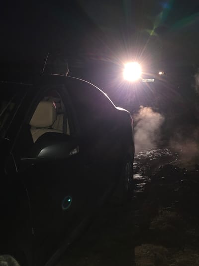 A dark, nighttime scene captures a vehicle with its headlights illuminating a muddy, uneven terrain. A person is partially visible in the driver's seat. White smoke or steam emanates from the area behind the vehicle, creating an atmospheric haze. The vehicle's license plate, P 719 TO 746, is clearly visible. The location is identified as Tagashevo, Russia.