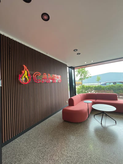 This image depicts a modern, unoccupied interior lounge or reception area located in Oostham, Belgium. The scene is dominated by a vertical slatted wooden accent wall on the left, featuring a vibrant neon sign. The sign consists of a stylized flame design in red and yellow neon, followed by the word "CAMPS" rendered in red neon, all mounted on a clear backing panel.

To the right, a large, frameless window offers an exterior view. Inside, a large L-shaped modular sofa, upholstered in a terracotta or burnt orange fabric with rounded edges, is positioned adjacent to the window. Two small tables accompany the sofa: a dark round side table with a slender black pedestal base, nestled into the sofa's corner, and a larger oval coffee table with a light gray speckled top and a black three-legged base, situated in front of the sofa.

The flooring throughout the interior is a light gray, speckled terrazzo or concrete. The white ceiling is fitted with multiple recessed circular light fixtures with dark bezels; some appear to be illuminated.

Through the large window, an outdoor environment is visible under an overcast sky. Green foliage, including bushes and small trees, lines the immediate exterior. Beyond the greenery, a paved area with several parked vehicles, including a dark blue sedan, can be observed. In the distance, a large, light blue or gray industrial building is prominently featured. No individuals are present in the image.