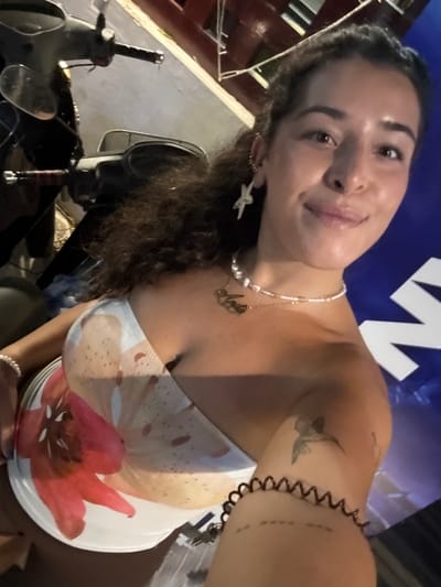 The image is a selfie taken by a young woman outdoors at night. She is leaning towards the camera, with her head tilted. The background is dark, with a large blue sign featuring white lettering partially visible. She is wearing a strapless top with a floral print. Around her neck, she has a pearl necklace with a gold pendant that appears to spell out "Anita". On her left ear, she wears a star-shaped earring. She has a tattoo of a bird on her upper right arm, and a coiled black bracelet on her wrist.
To the left of the woman, a motorcycle is visible. Parts of its handlebars and a mirror are in focus. The setting appears to be urban, consistent with the location context of Khan Daun Penh, Cambodia. The lighting suggests it is evening or night. The woman has a relaxed and happy expression, with a slight smile.
