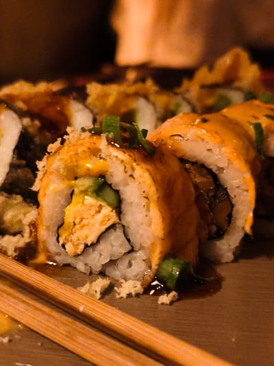 This is a close-up, dimly lit, shallow depth-of-field shot of several pieces of sushi on a wooden surface. The sushi rolls are topped with an orange sauce, possibly spicy mayonnaise or a similar creamy dressing, and garnished with finely chopped green onions. The sushi appears to be fried or tempura-battered, indicated by the crispy texture on some of the outer edges.
The primary subjects are the sushi rolls, presented in a close-up view that emphasizes their texture and toppings. Two wooden chopsticks are visible in the foreground, suggesting the act of eating or preparing to eat the sushi. The background is blurred and dark, with hints of warm lighting suggesting an indoor setting, possibly a restaurant or home.
The overall mood is intimate and appetizing, focusing on the food itself. The lighting is warm and low, creating a cozy atmosphere, and could indicate an evening meal. There are no visible people or text in the image. The context provided places this scene in Be'er Sheva, Israel, but this information cannot be derived from the image itself.