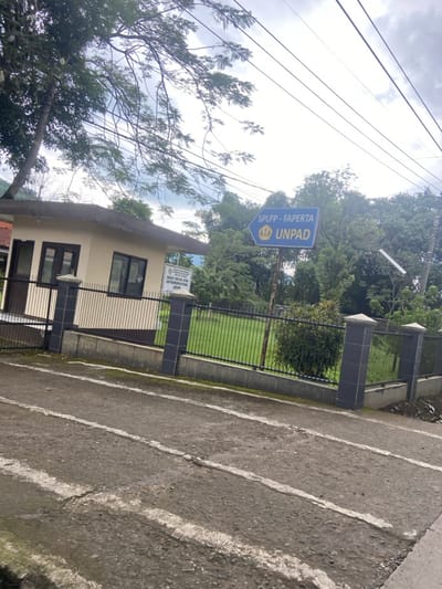 The image shows a paved road in front of a small, beige building with a black metal gate and fence.  The gate is closed. Behind the fence is a small, grassy yard. There is a blue sign above the gate, pointing right, which reads "SPLPP - FAPERTA UNPAD".  There are many trees and power lines overhead. The image was taken in Baleendah, Indonesia at the coordinates -7.022, 107.658. The scene is empty of people.  The ground is asphalt with white painted lines.